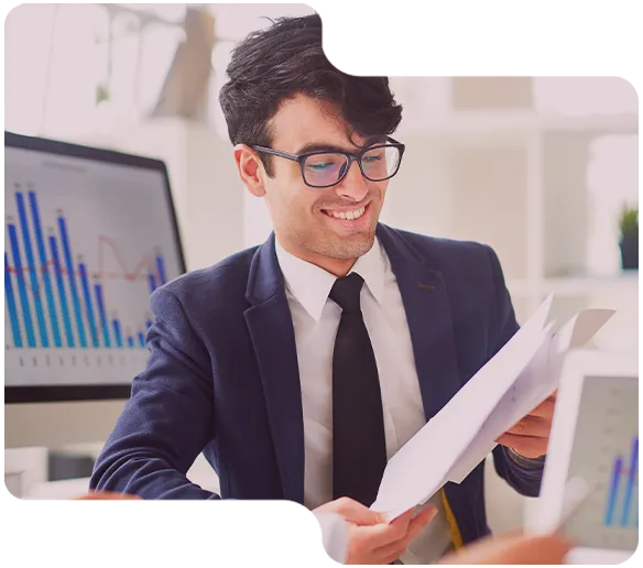 Smiling businessman in glasses reviewing documents with financial charts on computer screens nearby.