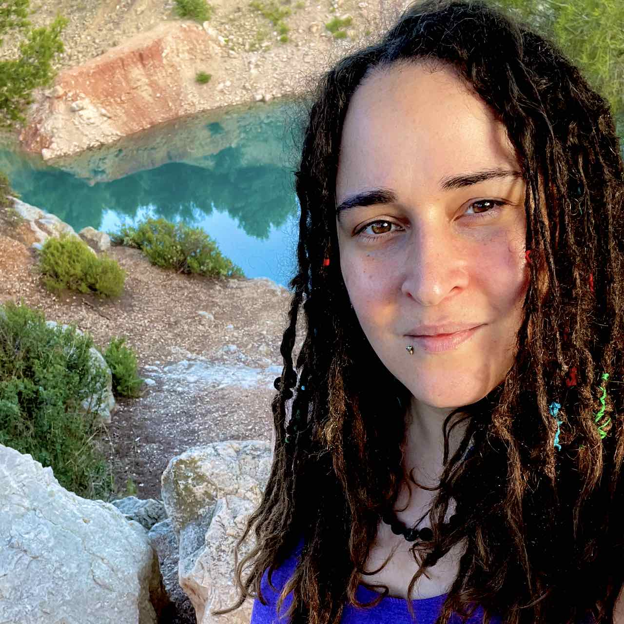 A selfie taken outside with a backdrop of various sizes of stones, conifers, an old bauxite quarry with some areas of red earth still visible & filled with turquoise water.