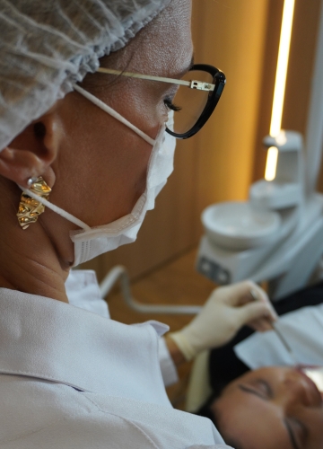 Profissional de saúde com máscara, touca e luvas realizando avaliação em paciente deitada.