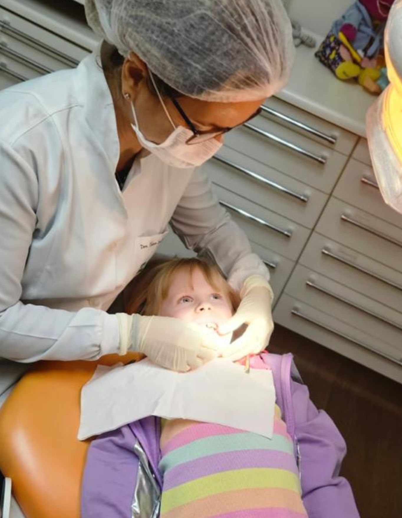Dentista com máscara e luvas examinando os dentes de uma criança na cadeira do consultório.