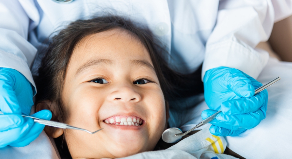 Criança sorrindo durante exame dentário com profissional usando luvas azuis e instrumentos odontológicos.
