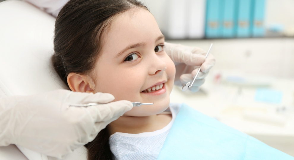 Criança sorridente sentada na cadeira do dentista com luvas e instrumentos próximos ao rosto.