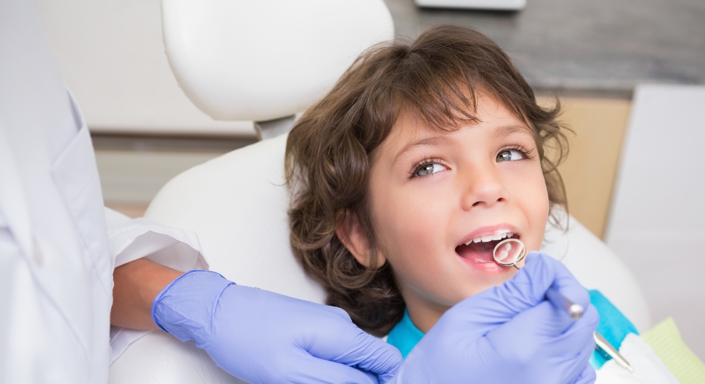 Dentista com luvas azuis examinando os dentes de uma criança sorridente na clínica.