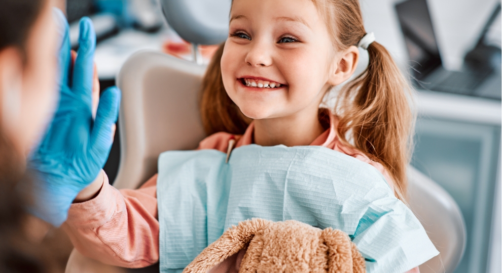 Menina sorridente no consultório odontológico fazendo high five com profissional de saúde.
