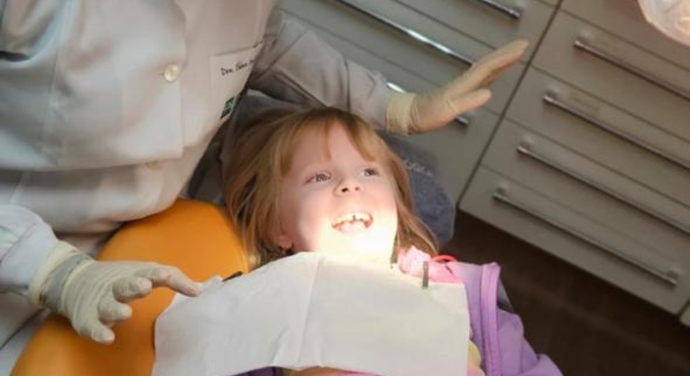 Criança sorridente sentada em cadeira de dentista durante exame odontológico.