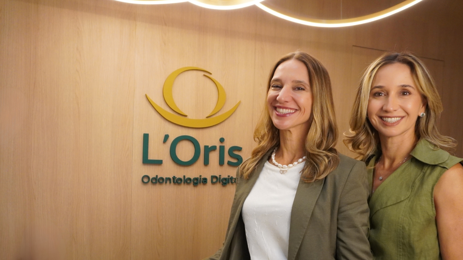 Duas mulheres sorrindo em frente a uma parede de madeira com o logotipo da clínica L'Oris Odontologia Digital.