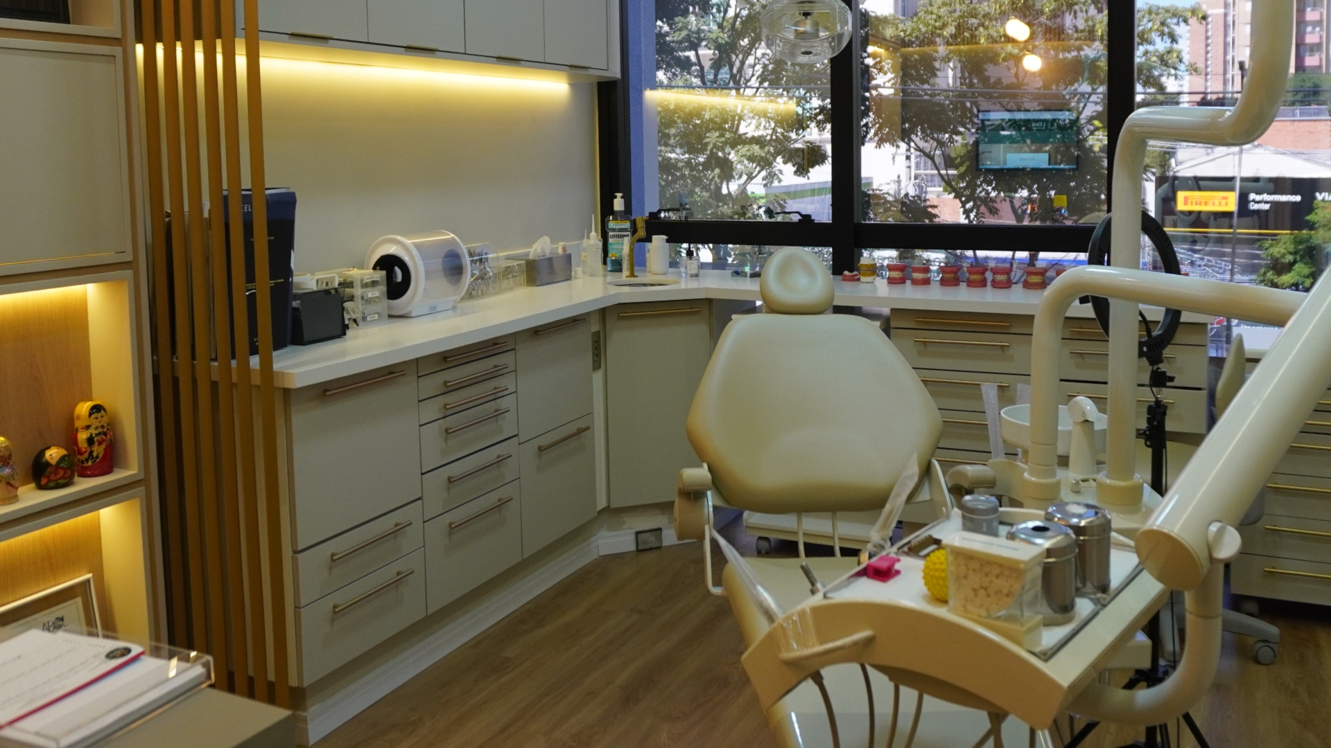 Consultório odontológico moderno com cadeira de dentista creme no centro, balcão em L com armários cinza claros ao redor, utensílios e modelos dentais em cima do balcão próximo à janela grande com vista para árvores e prédios.