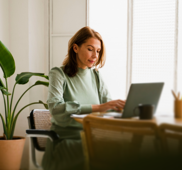 Woman in green dress typing on a laptop at a wooden table near a potted plant in a bright room.