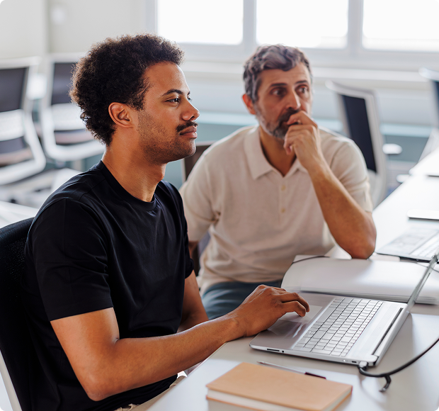 Two men working together at a desk looking at a laptop in a bright office setting.