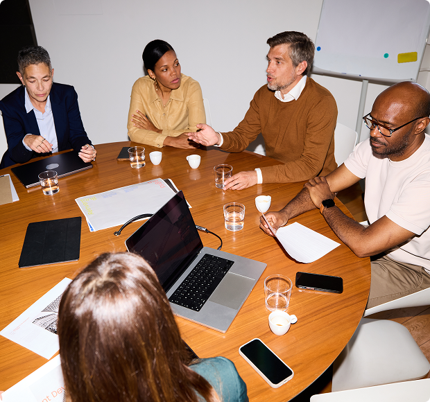 Five people having a discussion around a round table with laptops, papers, and drinks.