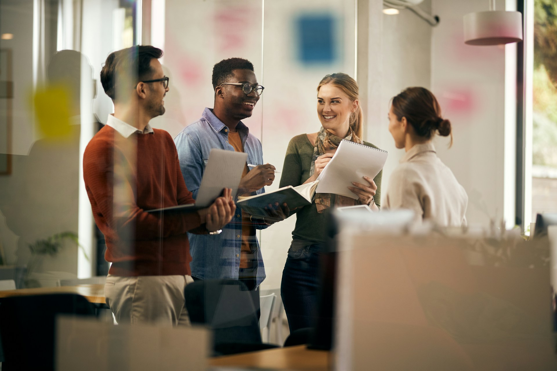 Four coworkers standing and discussing ideas while holding notebooks in a modern office.