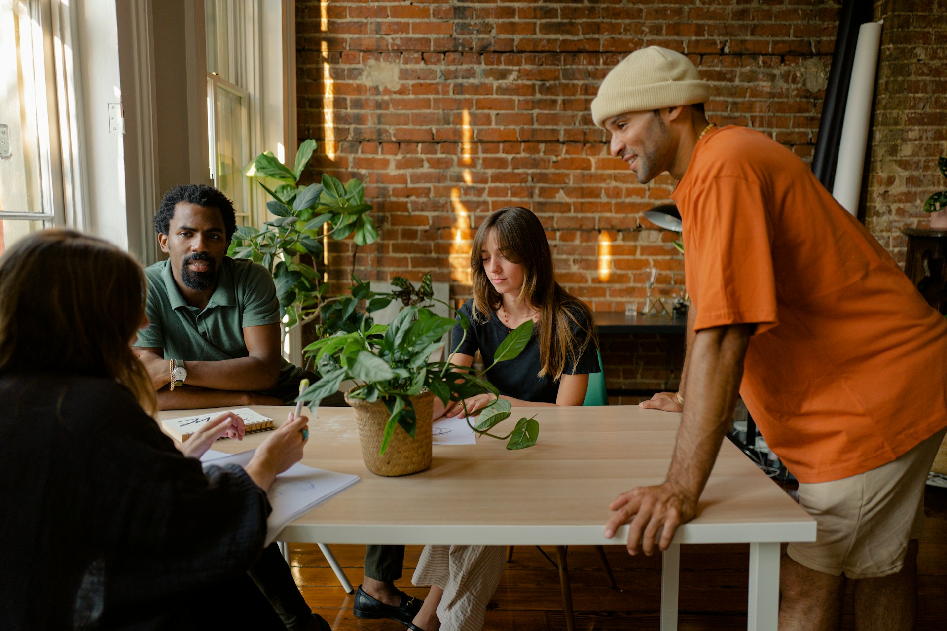 Four young adults collaborating at a table with plants, papers, and drawings in a casual office space.
