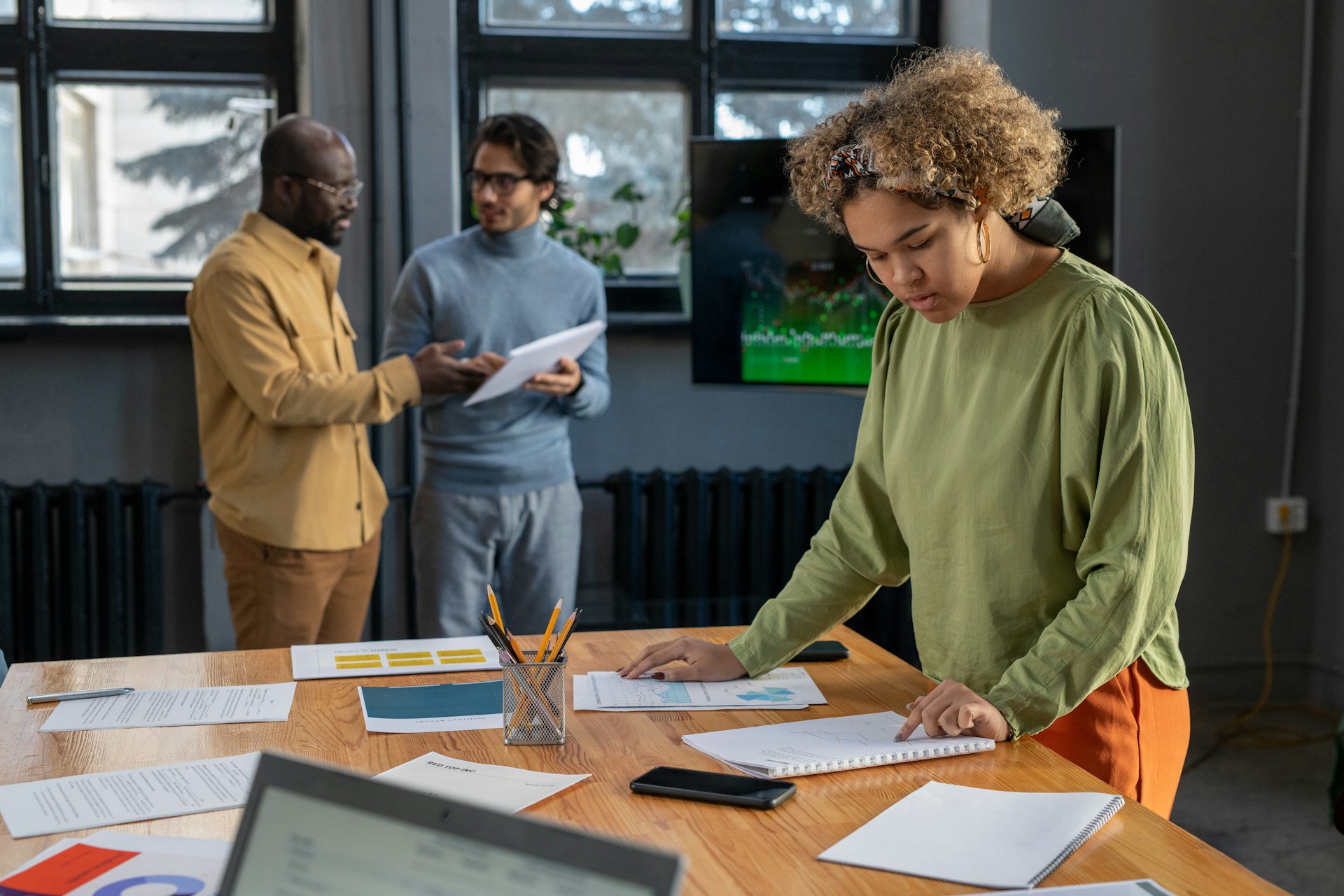 Woman in green shirt studies papers on table while two men discuss documents in background.