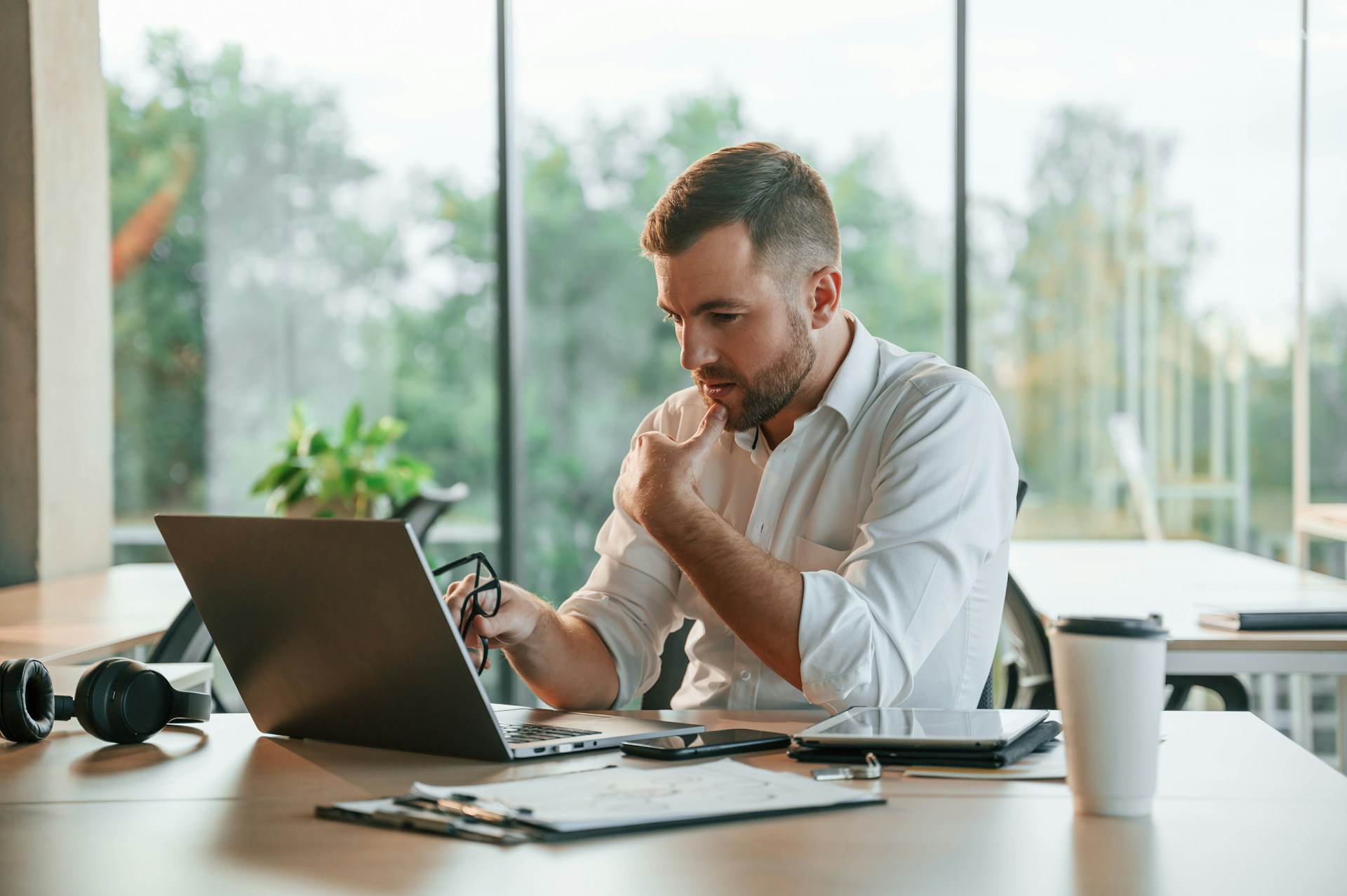 Man in white shirt looking thoughtfully at laptop while holding glasses in modern office.