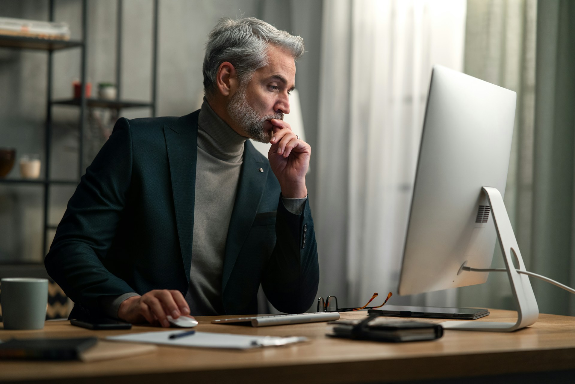 Mature man in blazer and turtleneck thoughtfully using computer mouse at desk with glasses and notepad.