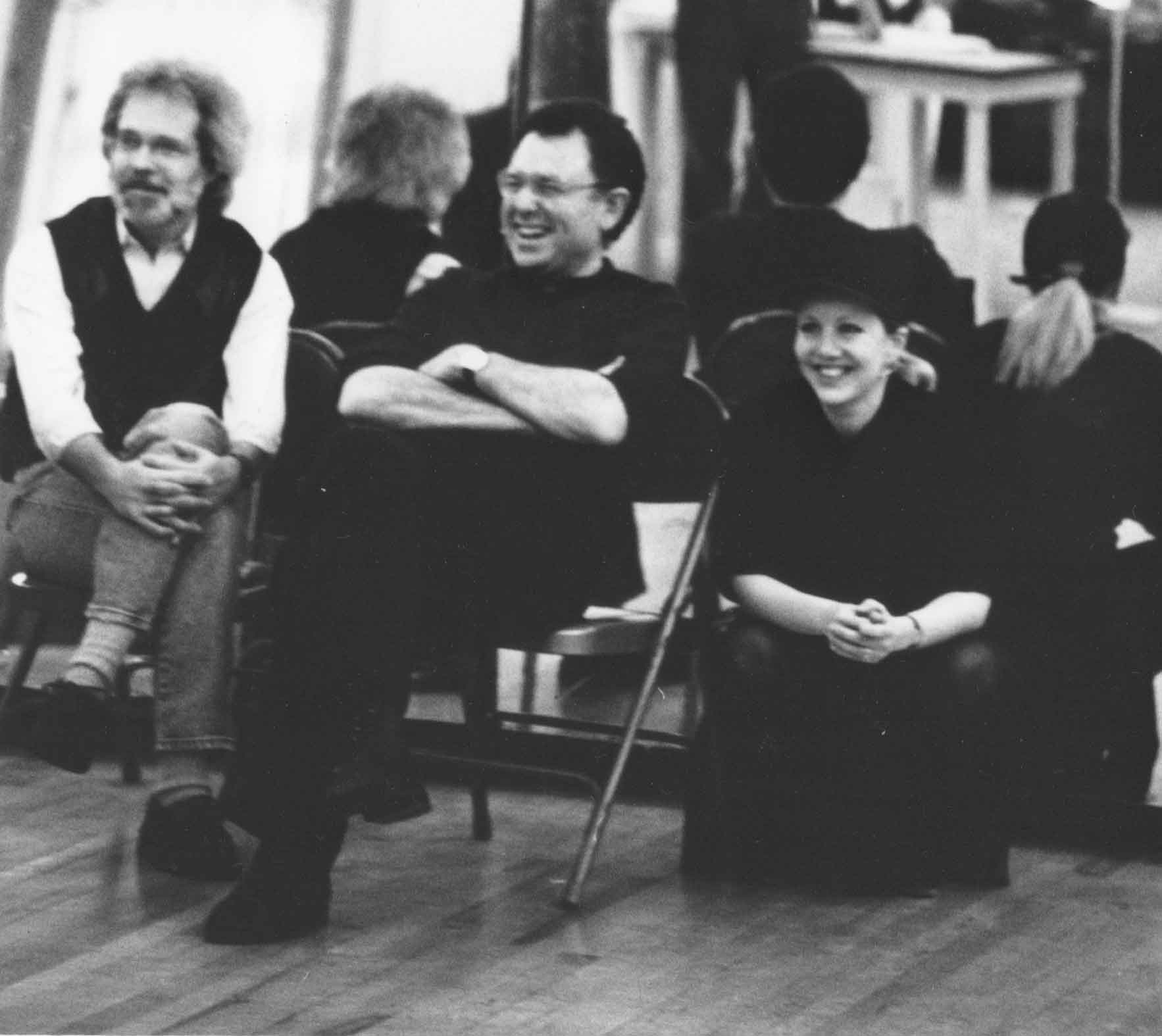 Three people sitting on chairs in a rehearsal space, smiling and relaxed, with a wooden floor.