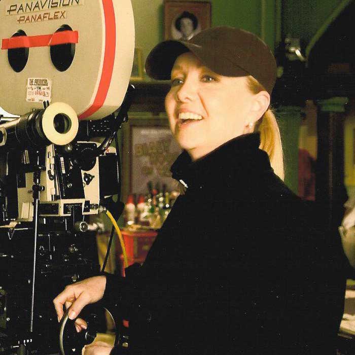 Woman in a black cap and jacket operating a Panavision Panaflex film camera indoors.