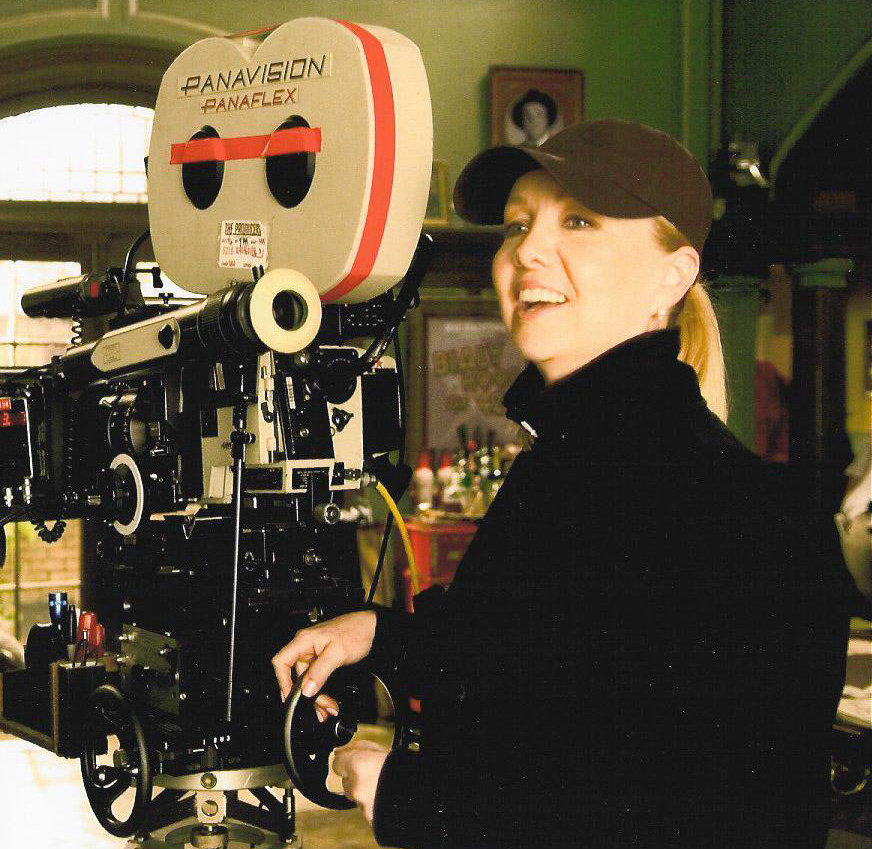 Woman in black jacket and cap operating a large Panavision Panaflex film camera on set.