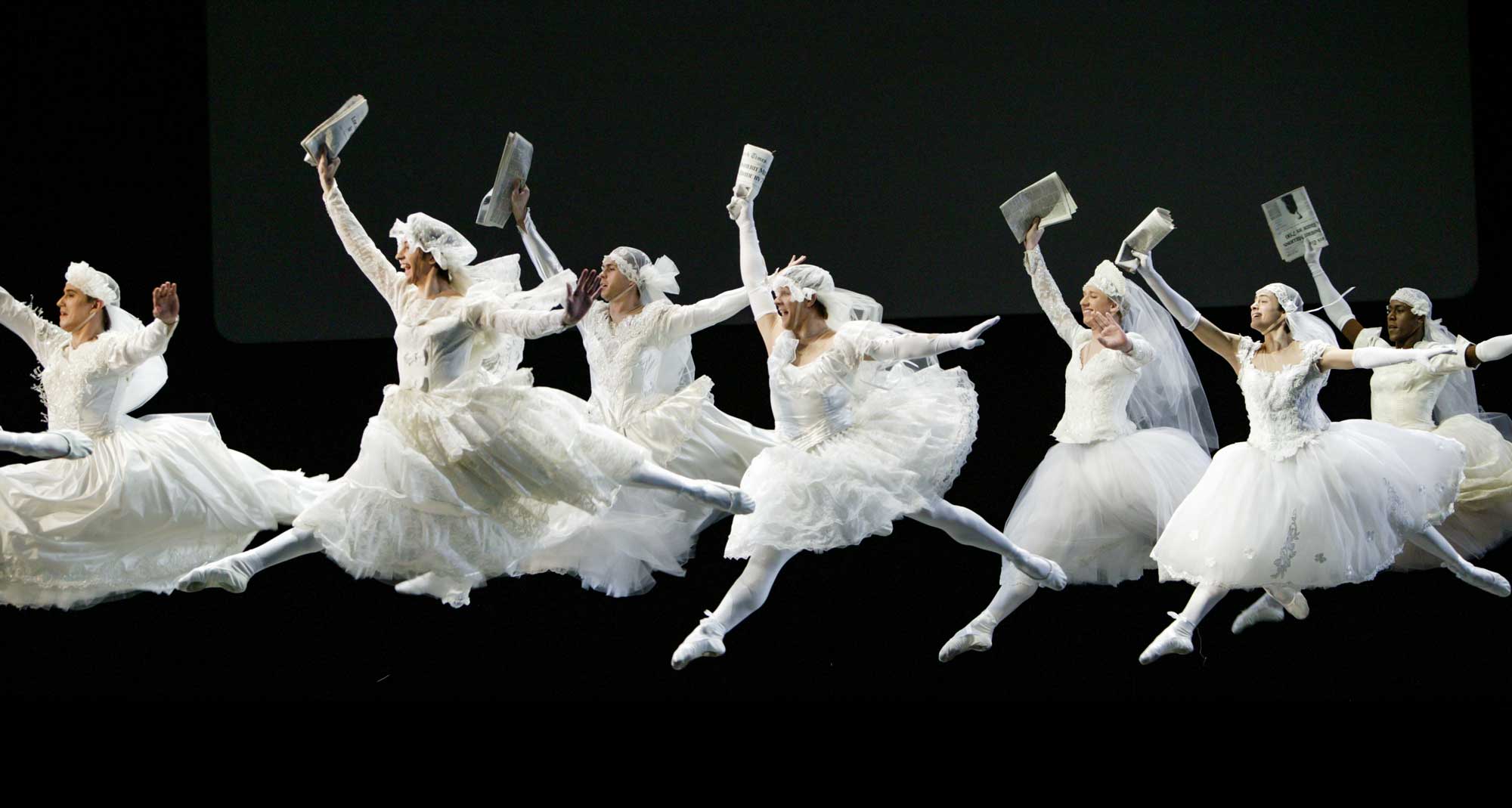 Group of ballet dancers dressed as brides in white tutus and veils, holding newspapers and performing grand jeté leaps on stage with a black background.