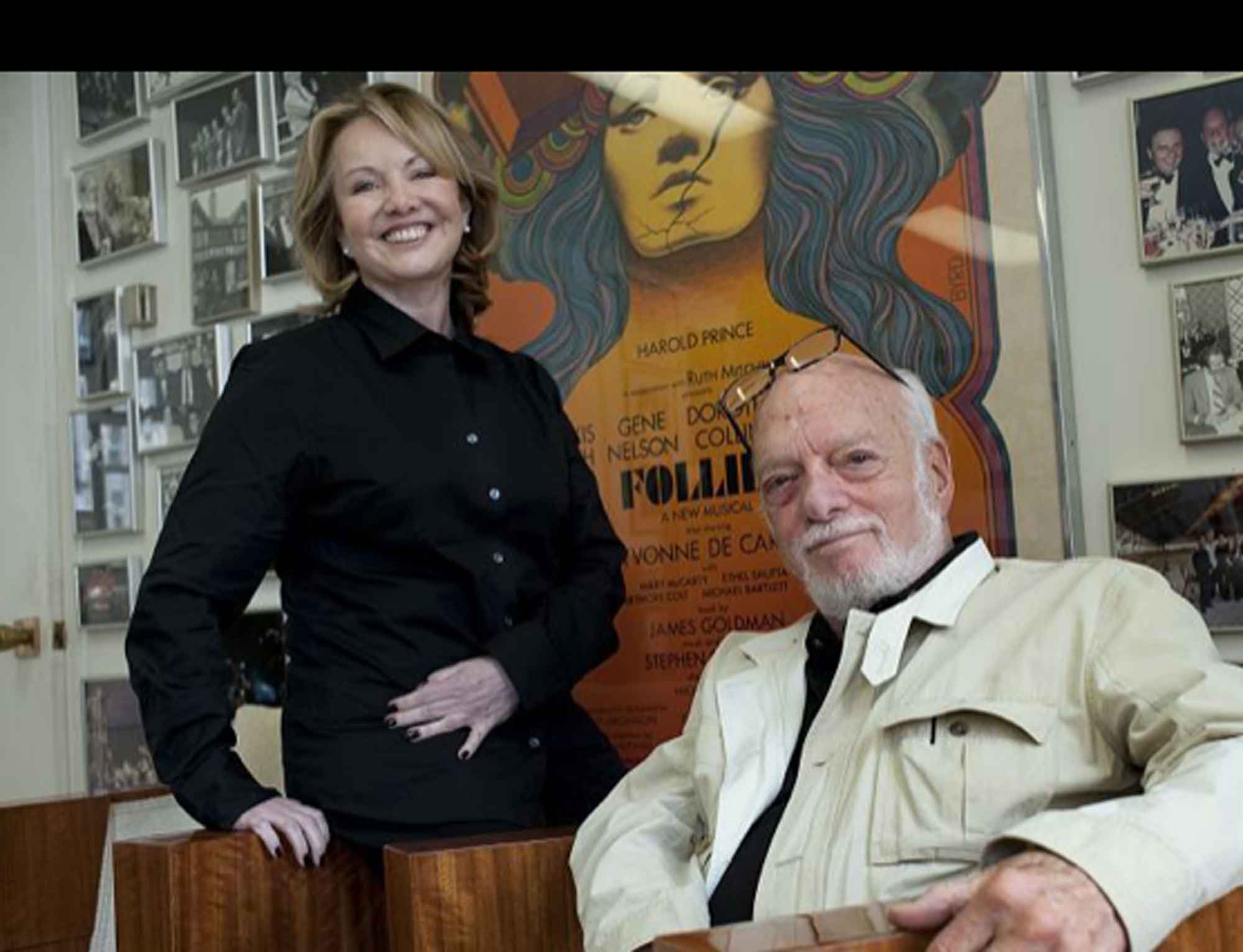 Smiling woman in black shirt standing next to seated older man in light jacket with glasses on his forehead, with a colorful poster and numerous framed photos on the wall behind them.