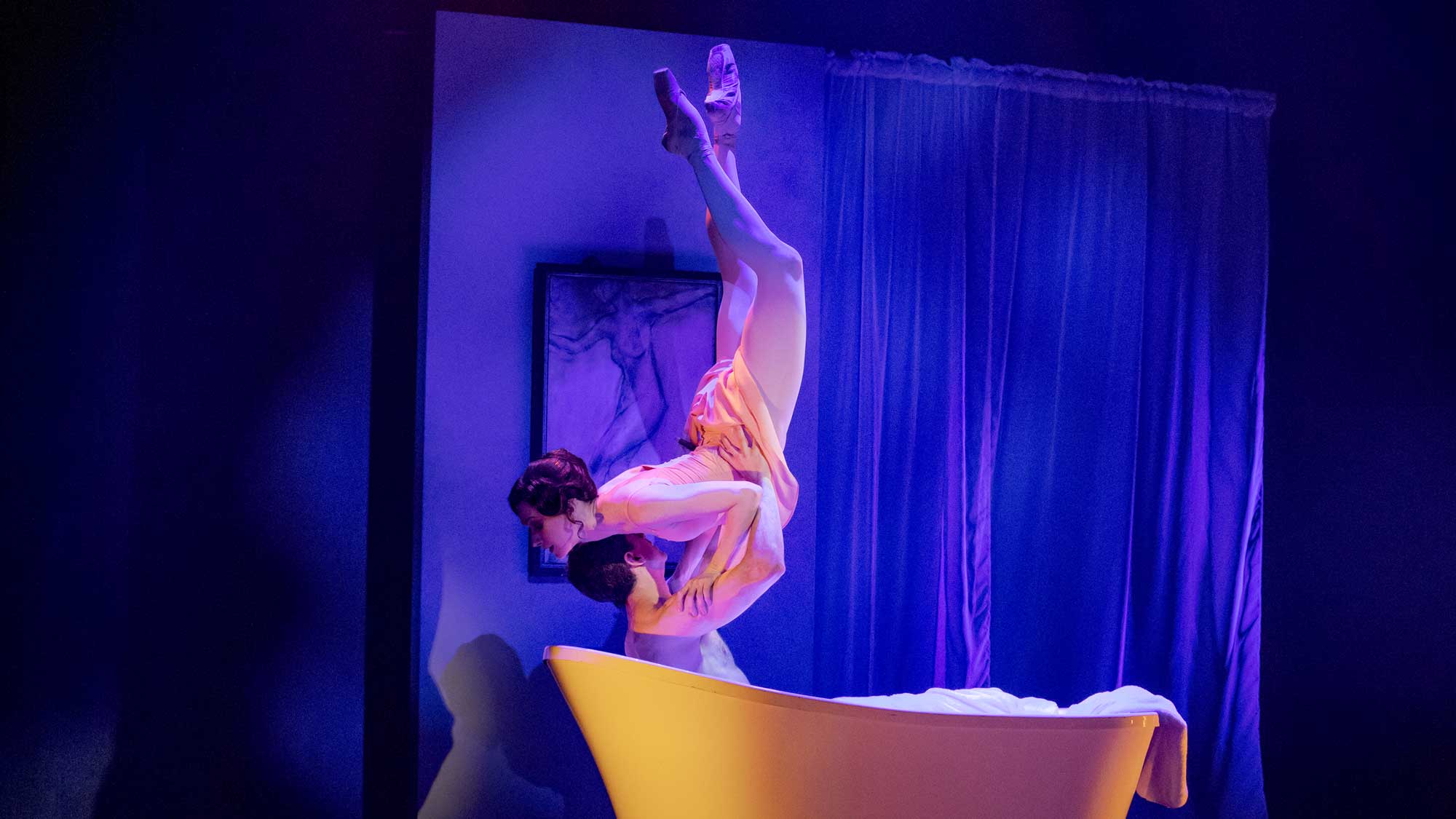 A male dancer lifts a female dancer upside down above a yellow bathtub on stage with blue and purple lighting.