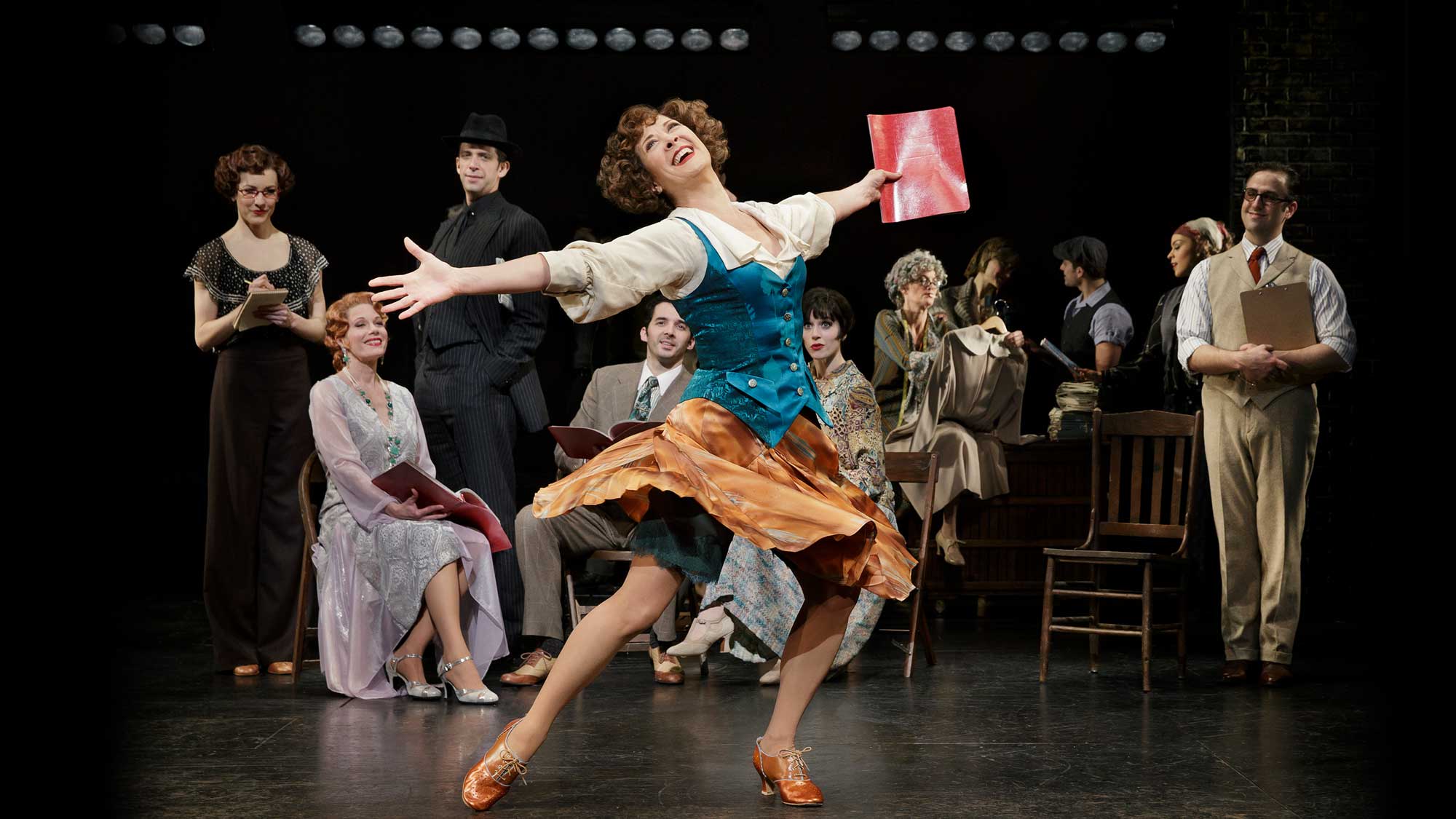 Actress in a vibrant skirt and teal vest joyfully dancing center stage with a red notebook, surrounded by seated and standing actors in vintage costumes.