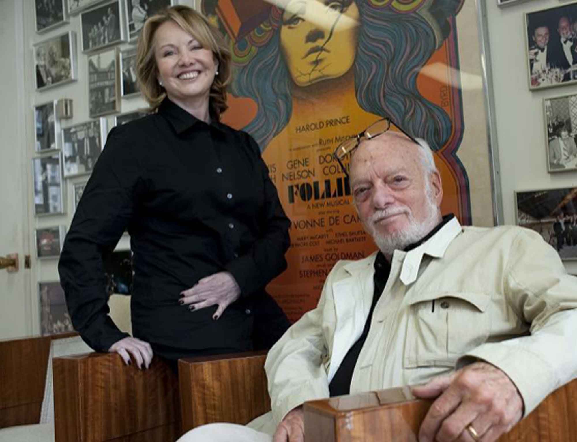 Smiling older man seated in a chair wearing a light jacket and a woman standing beside him in a black shirt, with a colorful poster and framed photographs on the wall behind them.