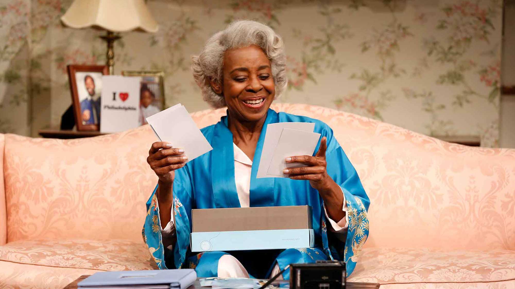 Smiling elderly woman wearing a blue robe sitting on a couch, sorting through printed photographs.