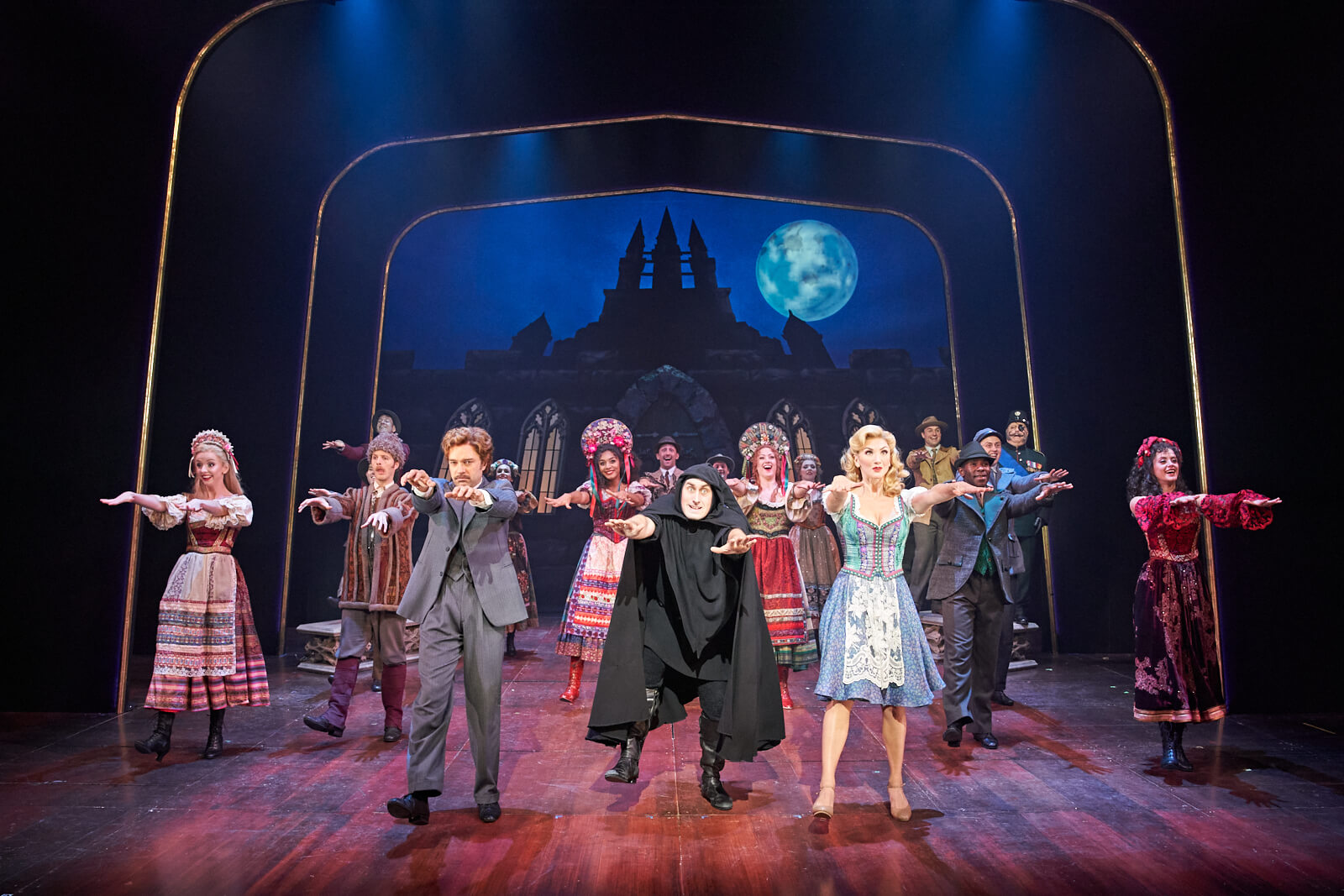 Dr. Frankenstein (Hadley Fraser), Igor (Ross Noble) and Inga (Summer Strallen) doing the monster step in "Transylvania Mania" with the happy villagers in front of the castle. 