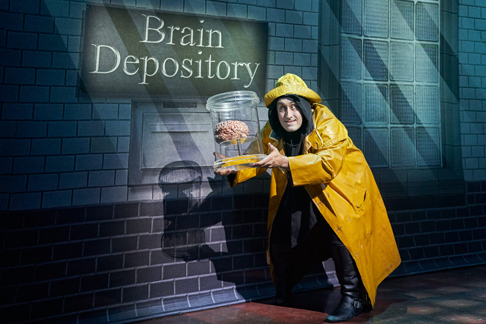 Igor (Ross Noble) in a yellow raincoat stealing a brain from a brain depository in Young Frankenstein (West End).