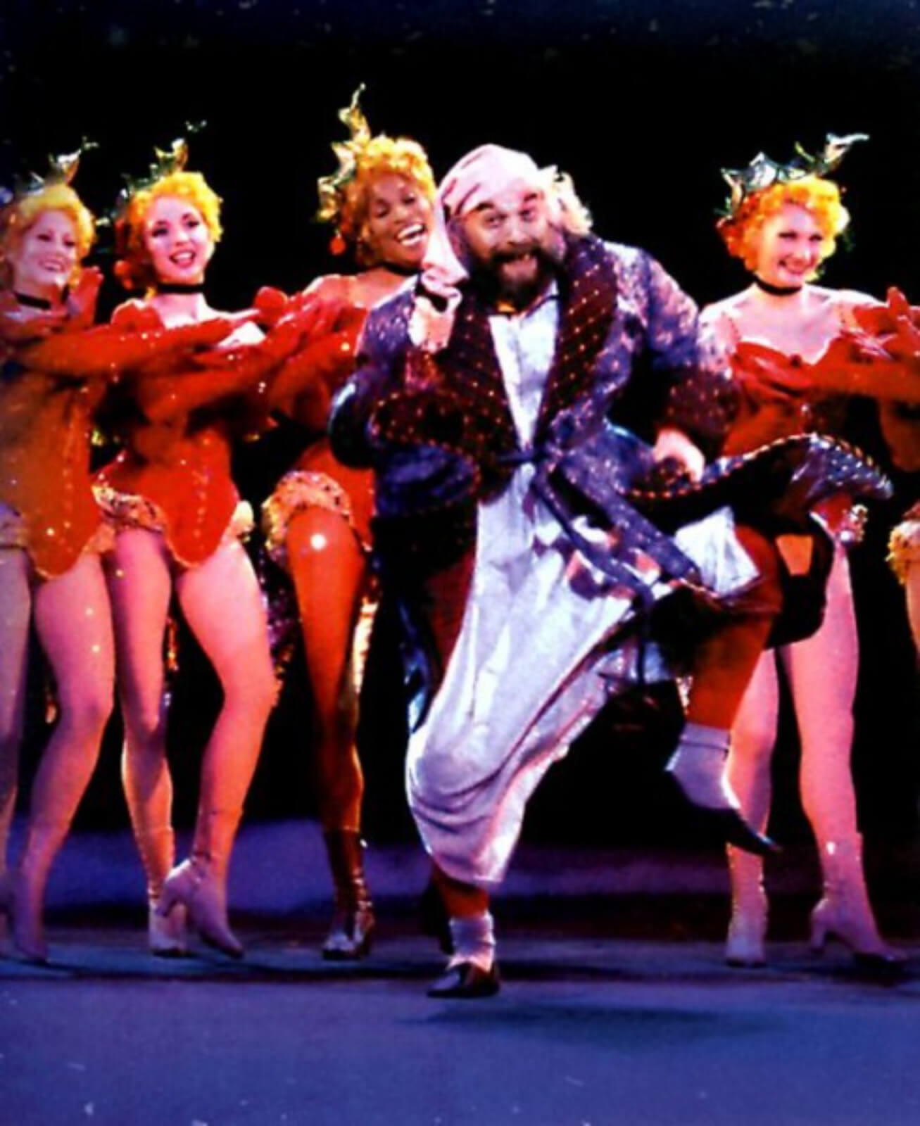 Scrooge (Walter Charles) in a robe happily dancing as he is surrounded by Holly Girls in bright red costumes