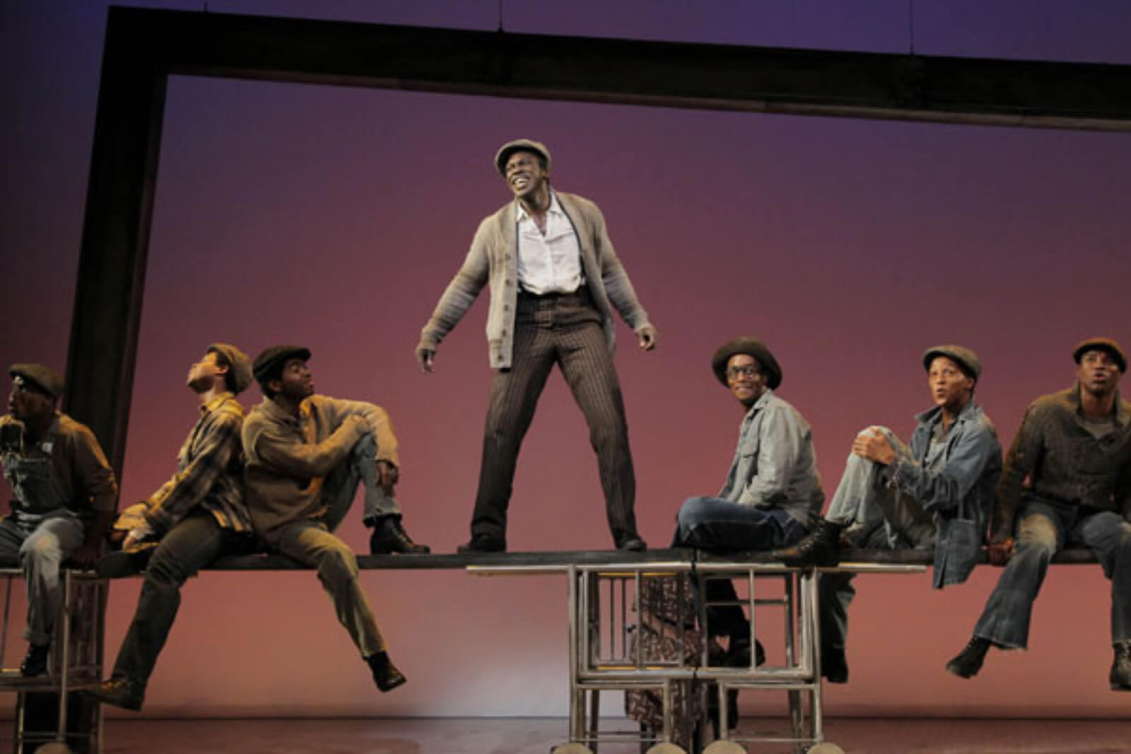 Haywood Patterson (Joshua Henry) and the Scottsboro Boys sing "Commencing in Chattanooga". They are balancing on beams that represent the top of a train. They are dressed in work clothes.