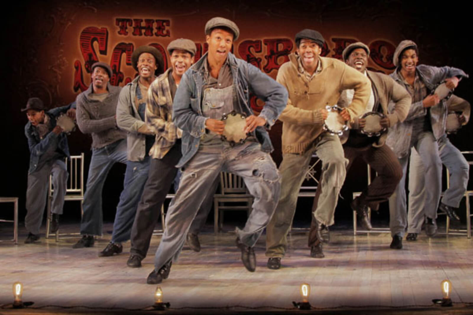 The Scottsboro Boys sing and dance in "Hey, Hey, Hey, Hey!" The actors are in a V-formation holding tambourines, doing a vaudeville flea hop step behind the footlights.