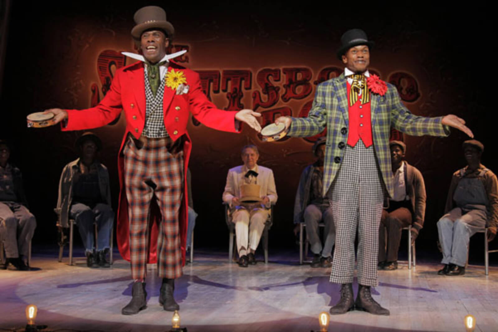 Mr. Bones (Coleman Domingo) and Mr. Tambo (Forrest McClendon) singing "Hey, Hey, Hey, Hey!" The title song in the show. They have tambourines in their hands and are wearing colorful costumes.