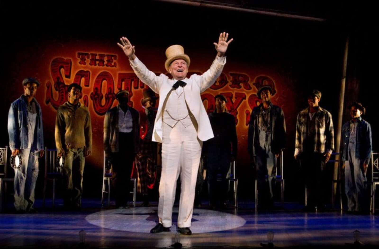 The Interlocutor (John Cullum) is saying "Gentleman be seated." He is wearing a white linen suit and a white top hat. He is in a hard edged spot. The Scottsboro Boys are in the background.