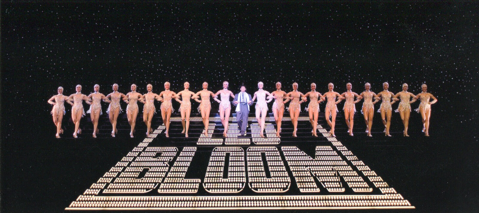 Leo Bloom (Matthew Broderick) and The Pearl Girls. They are walking down a large stair case with the word Leo Bloom painted in the center. There are 10 girls on each side of him. There is a star drop behind them.