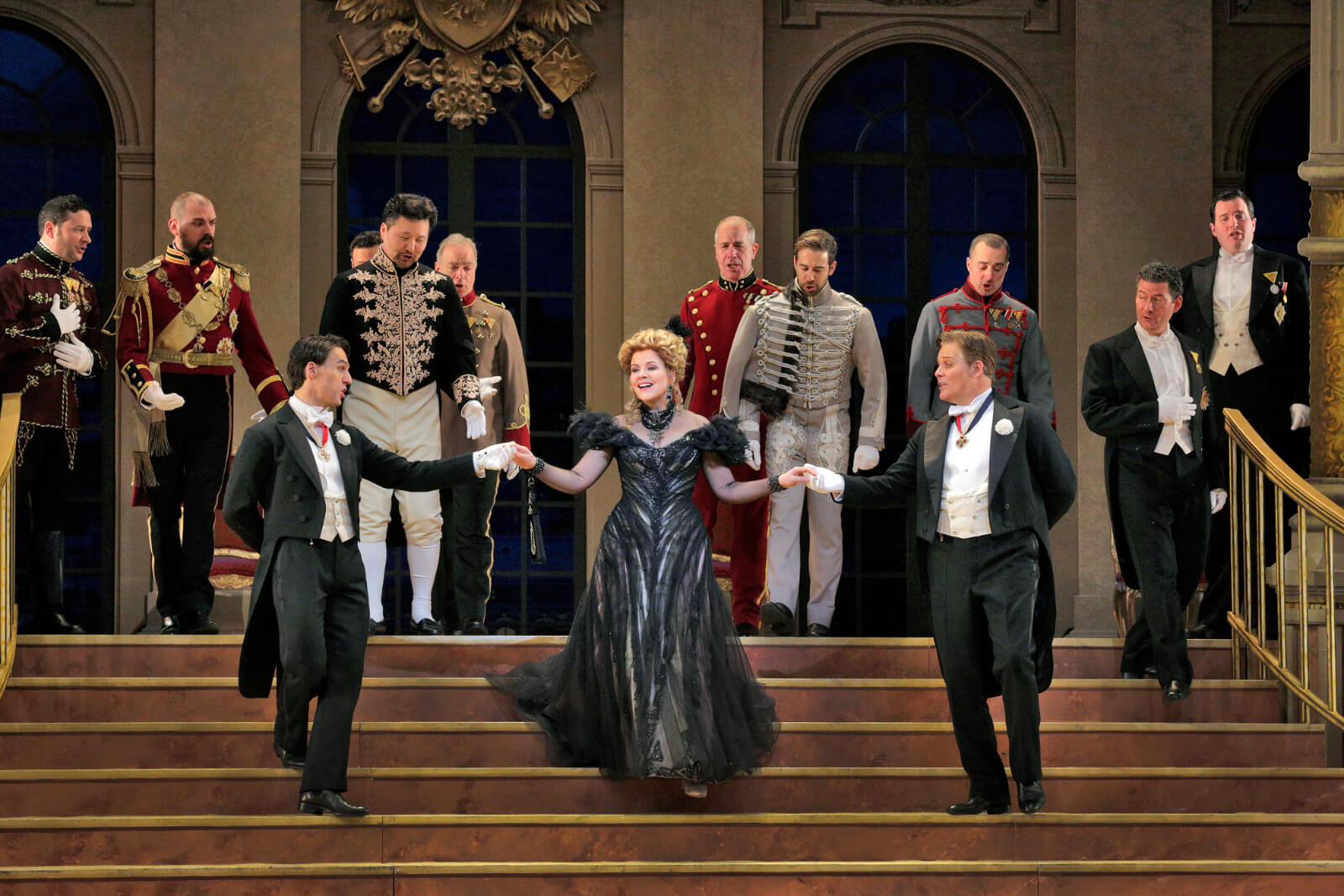 "Hanna's Entrance", with Renee Fleming and her gallant suitors in the Embassy Ball. She is wearing a black gown. Two gentlemen escort her down the stairs, surrounded by other suitors. 