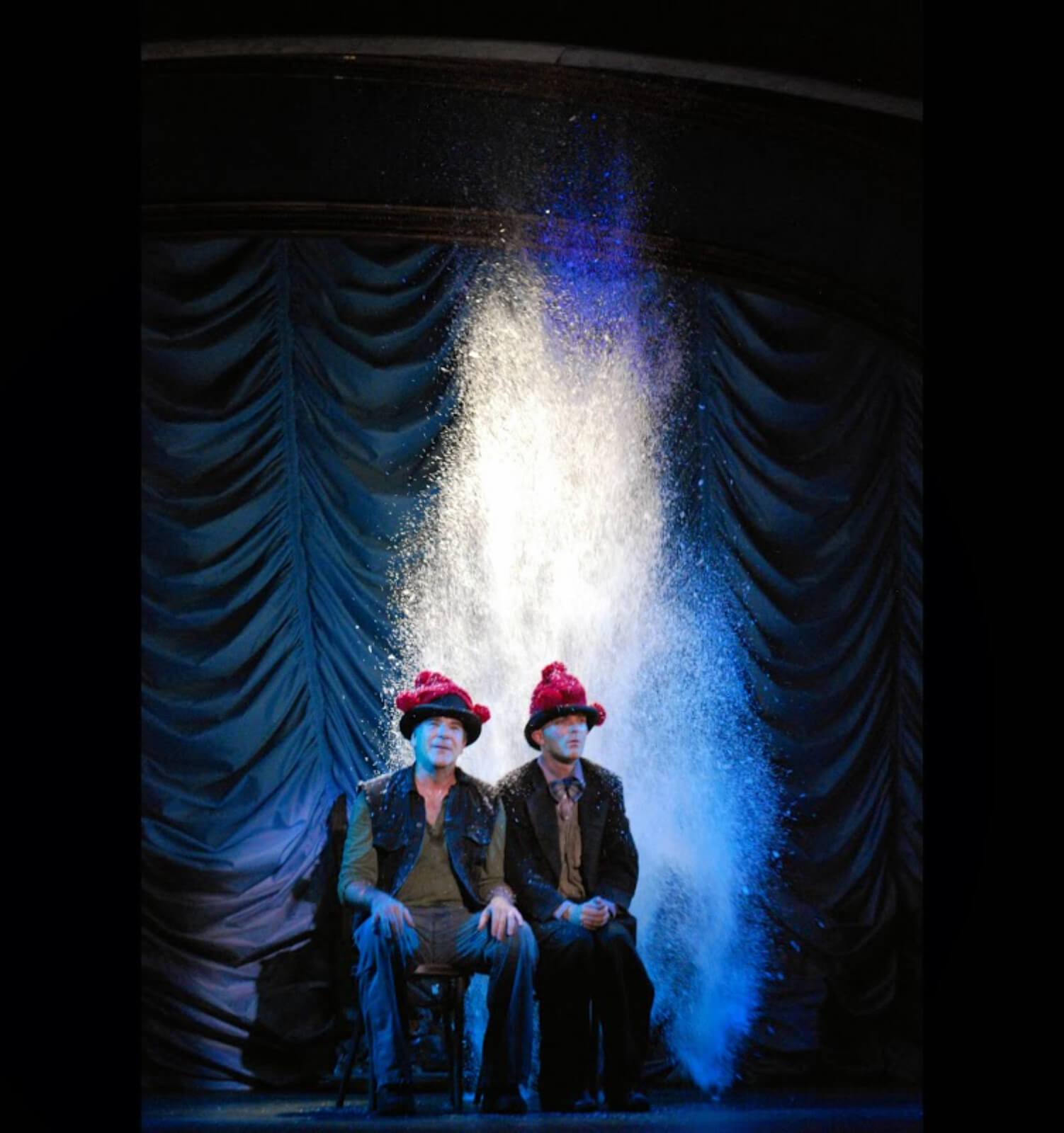 Mandy Patinkin and Taylor Mac singing "Snow". They are sitting close together with red wool hats on and a large amount of snow is falling from above. 