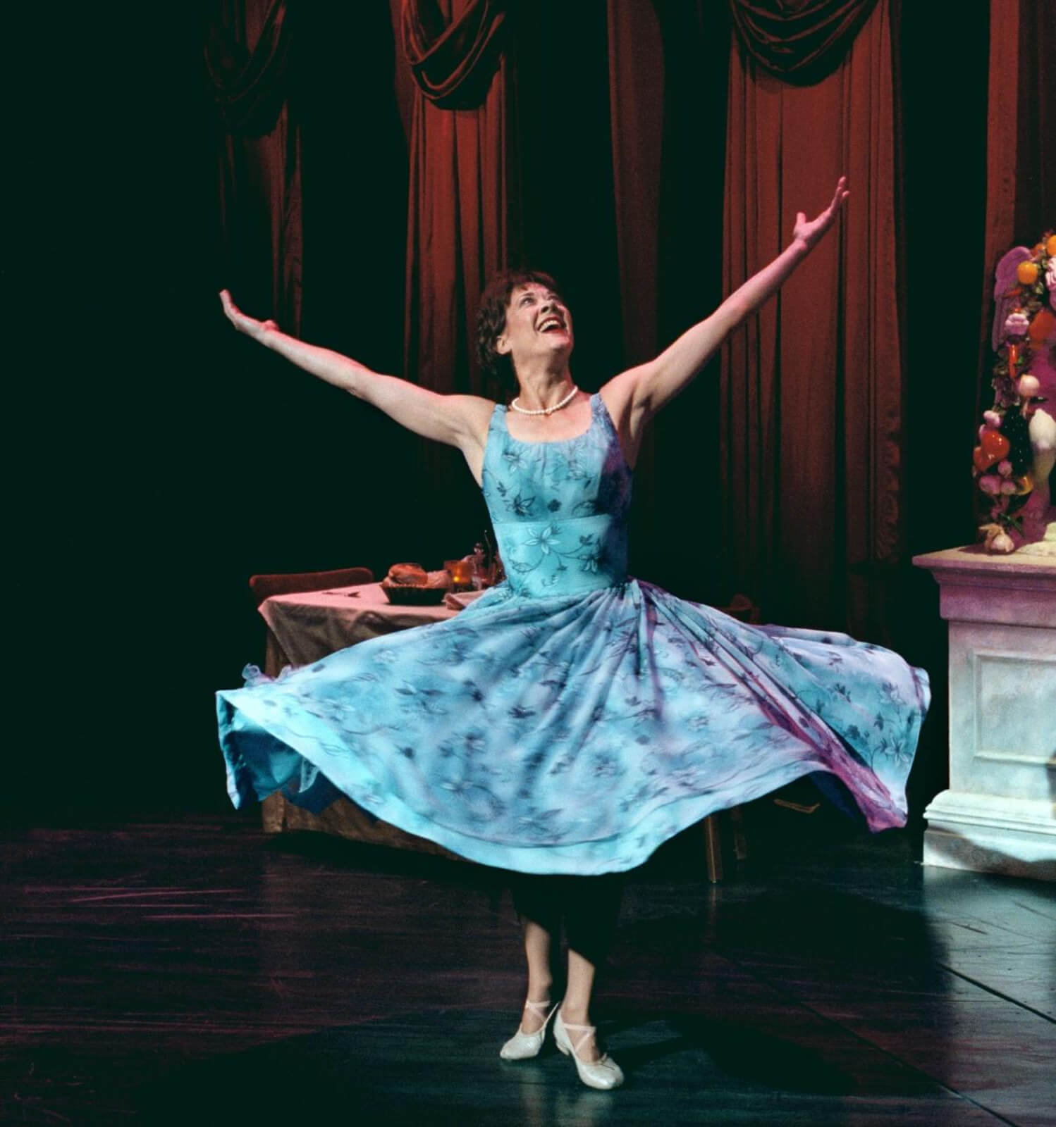 Wife (Karen Ziemba) twirling with her arms up in a restaurant. She is wearing a beautiful blue dress in Contact.