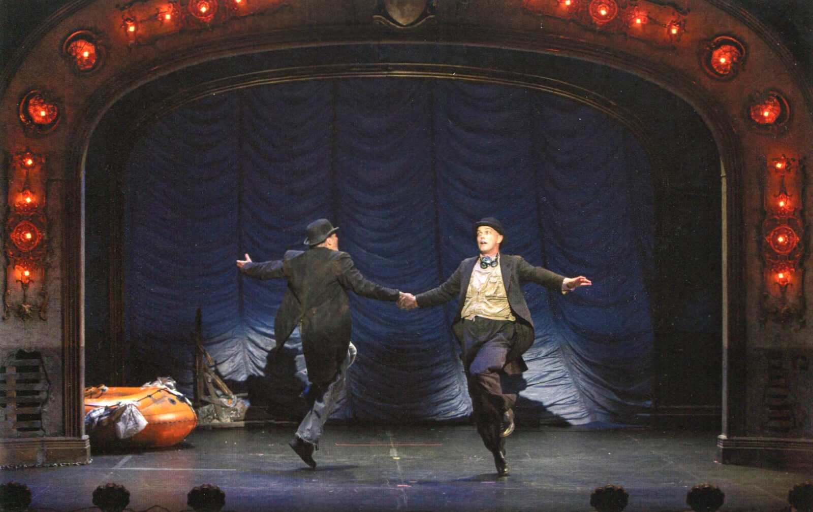 Mandy Patinkin and Taylor Mac dancing to "It's the End of the World As We Know It". They are skipping and holding their right hands together. 