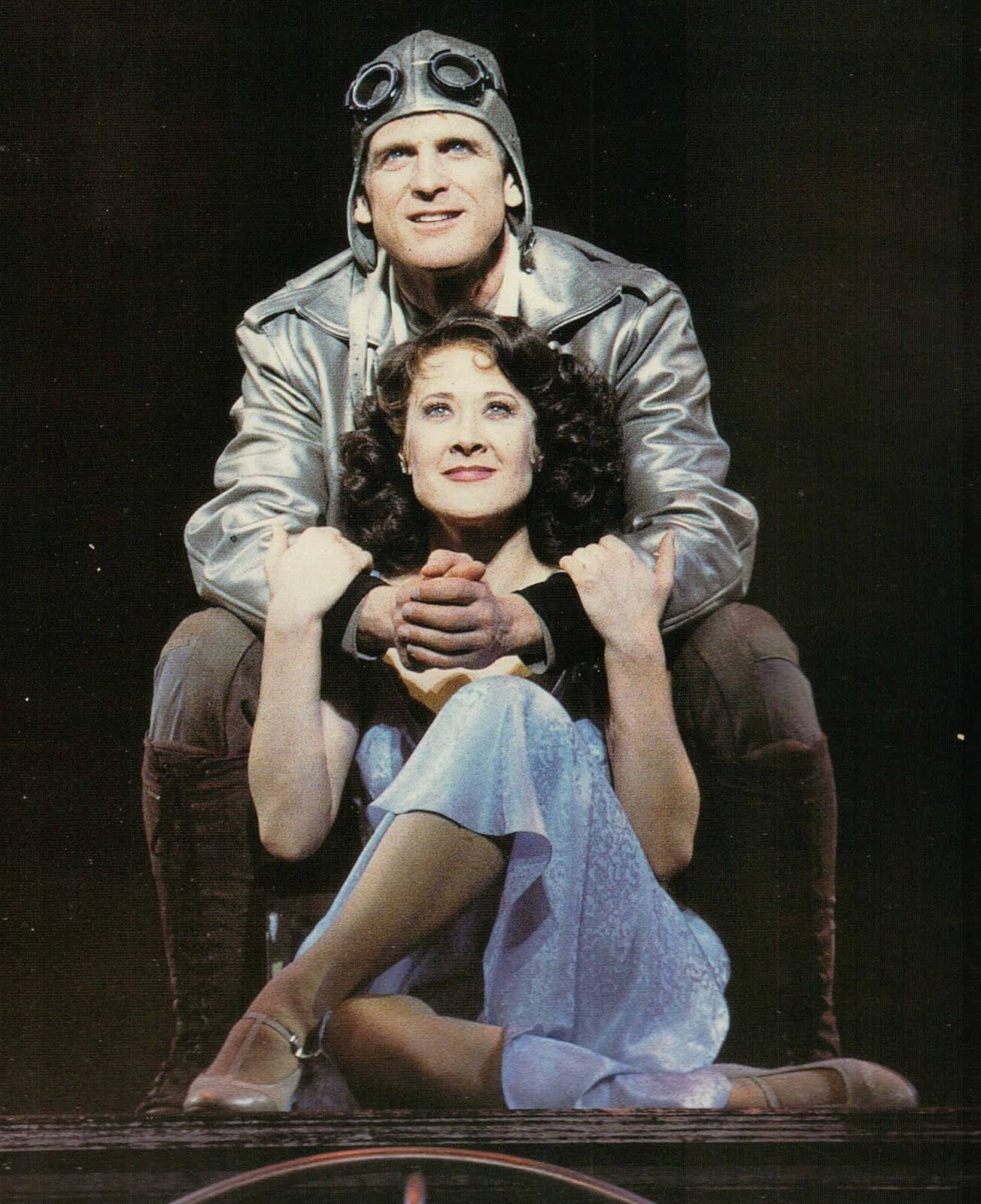 Bill Kelly (Daniel McDonald) in a pilot suit, embracing Rita Racine (Karen Ziemba) from behind. Rita holding his arms singing "First You Dream".