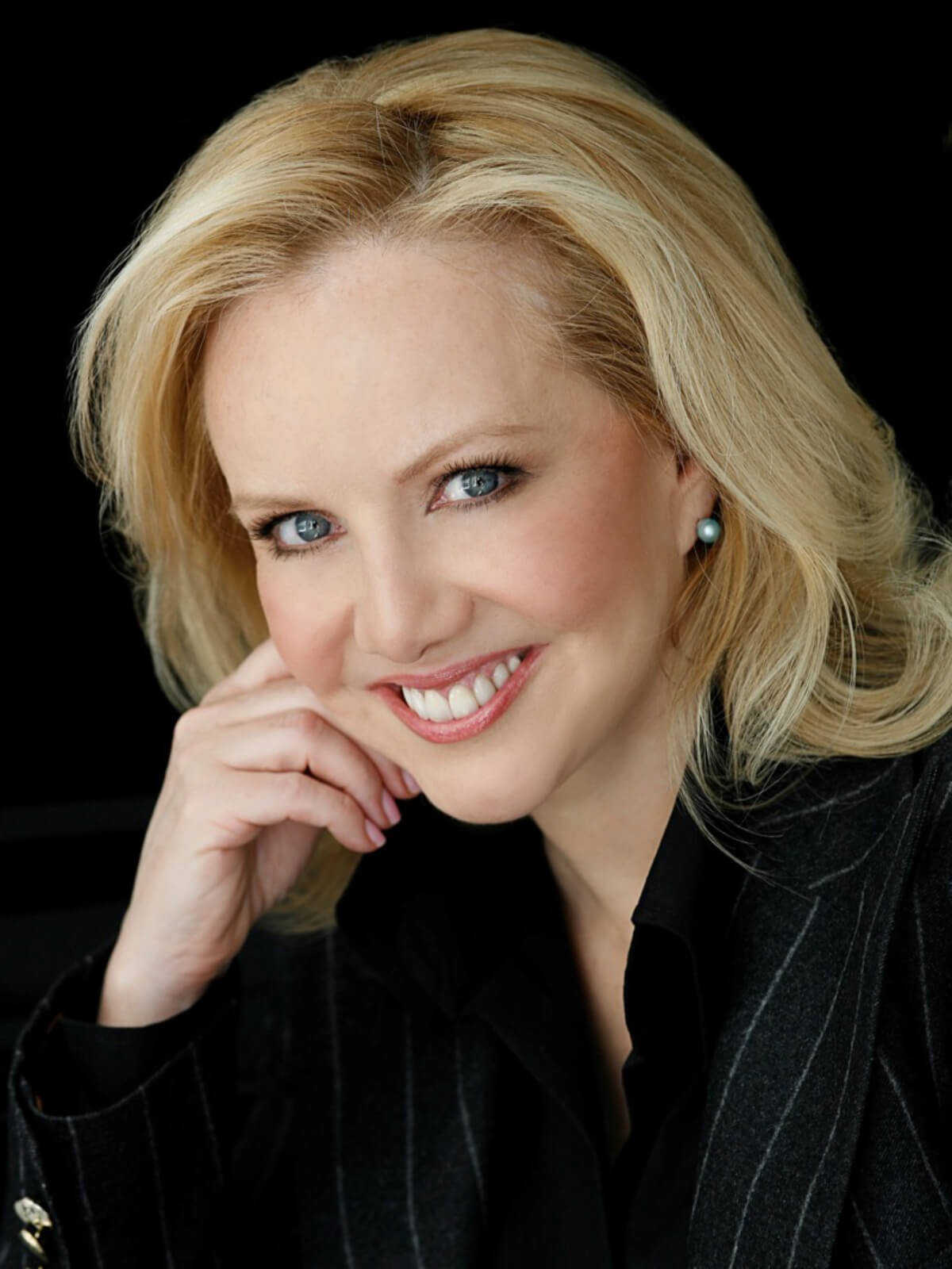 Headshot of Susan Stroman. She is wearing a pinstriped black suit smiling to camera with head resting on upstage hand.