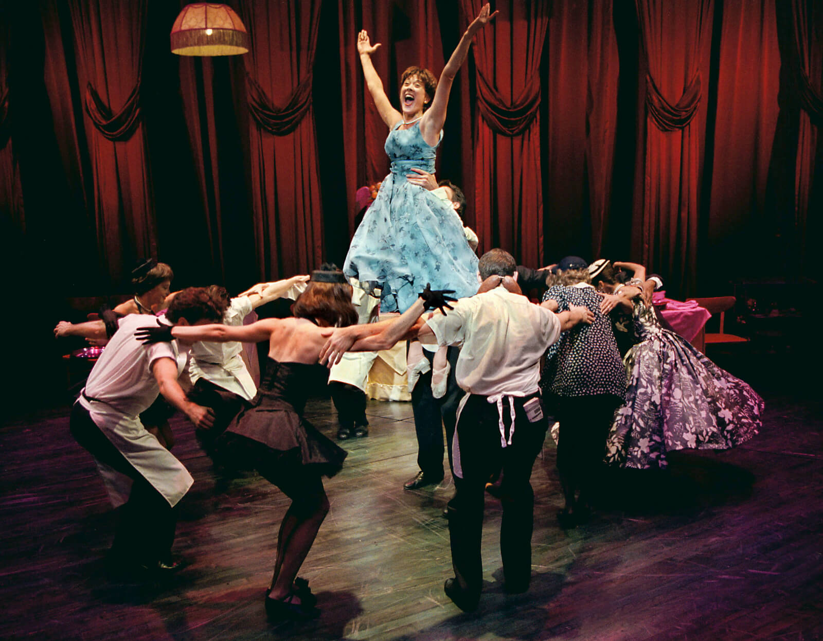 Wife (Karen Ziemba) wearing a blue dress is lifted with outstretched arms while surrounded by the full company.