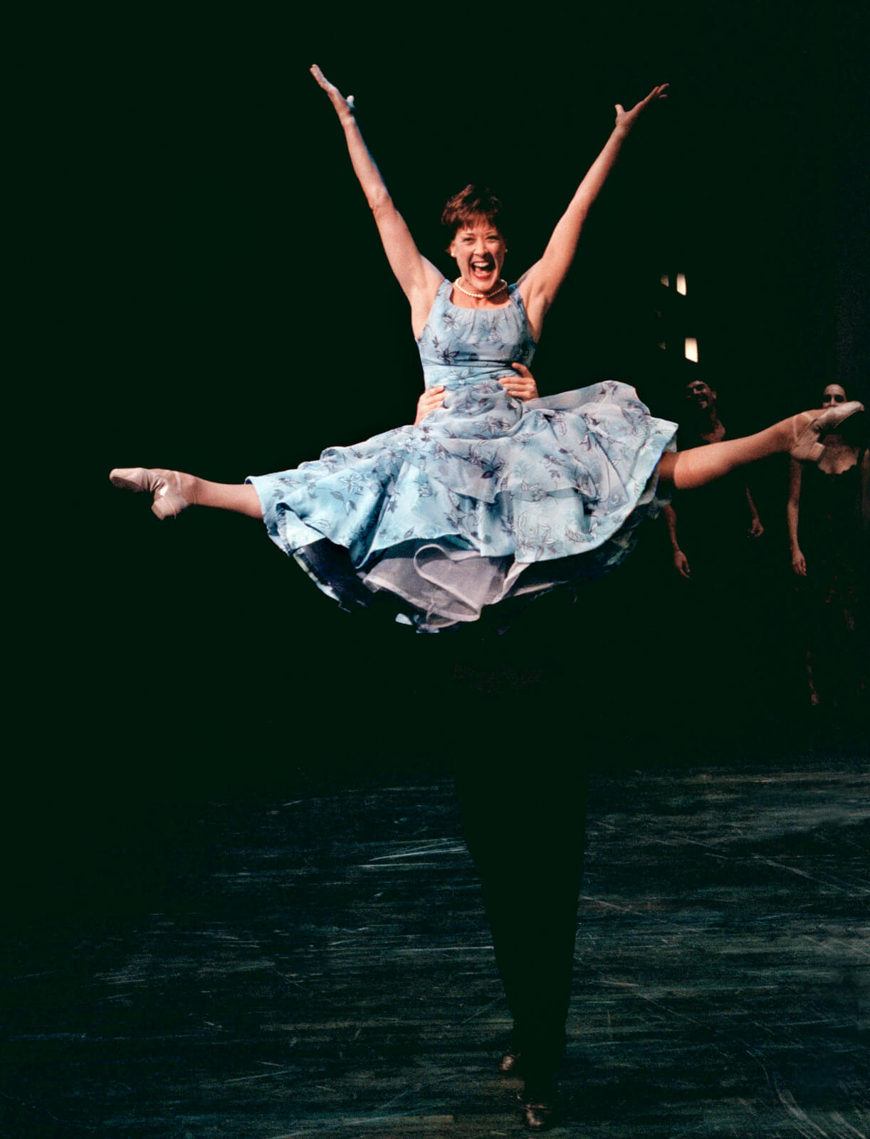 Wife (Karen Ziemba) in a blue dress is lifted into the air in a full second position split. 