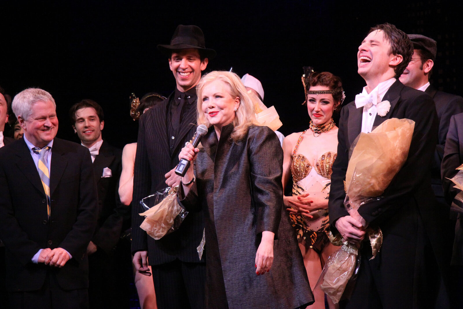 Susan Stroman at the opening night curtain call of Bullets Over Broadway, standing with a microphone and flanked by cast, including Nick Cordero and Zach Braff.