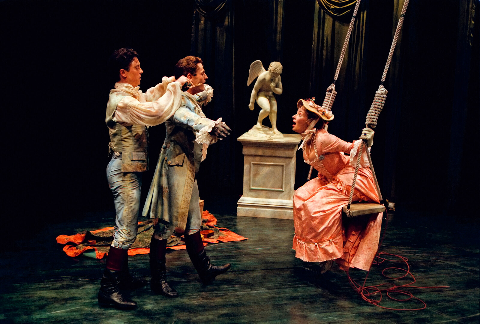 Scott Taylor, Seán Martin Hingston approaching Stephanie Michels on the swing. She is looking at them with a shocked expression. The young woman is on the swing with a billowing pink skirt, Scott Taylor the Servant is putting the jacket onto the husband.