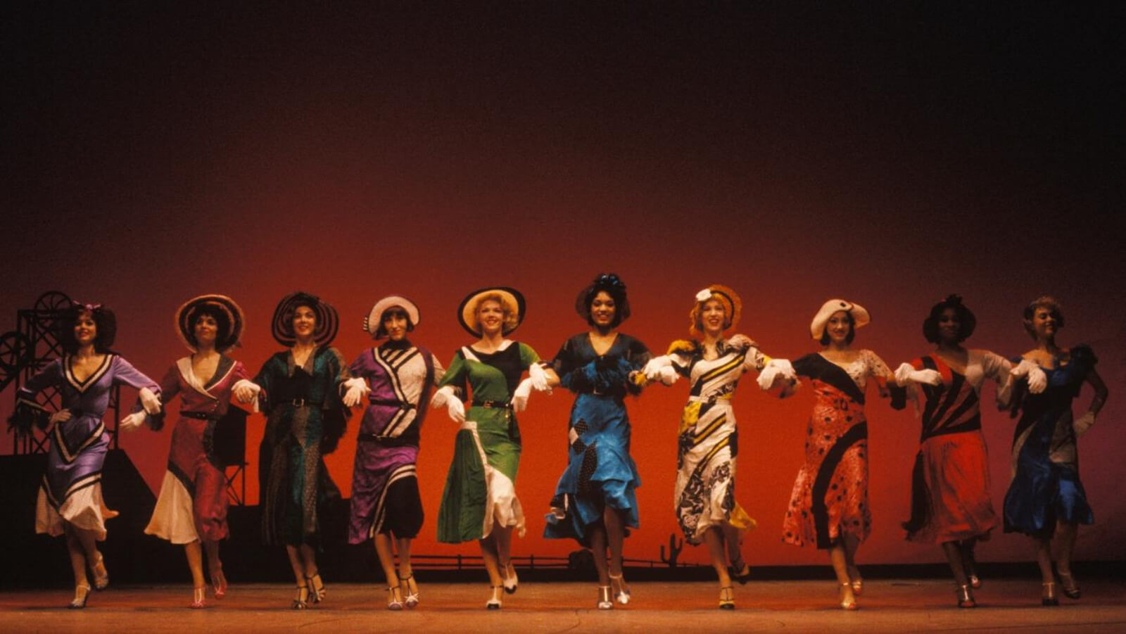 The Zangler Follies girls make their entrance into the town of Deadrock, Nevada in Crazy for You. The Zangler Follies are dressed in brightly colored dresses and hats as they link arms and arrive in the town of Deadrock