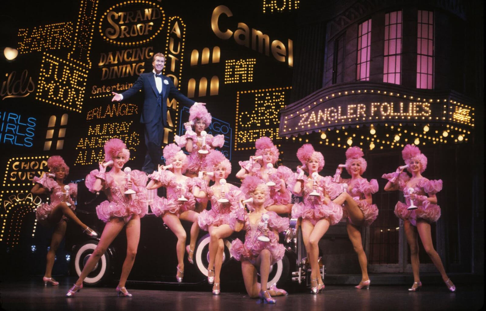 Bobby Child (Harry Groener) stands on top of a car with 10 female dancers all in matching pink tutus posed around him