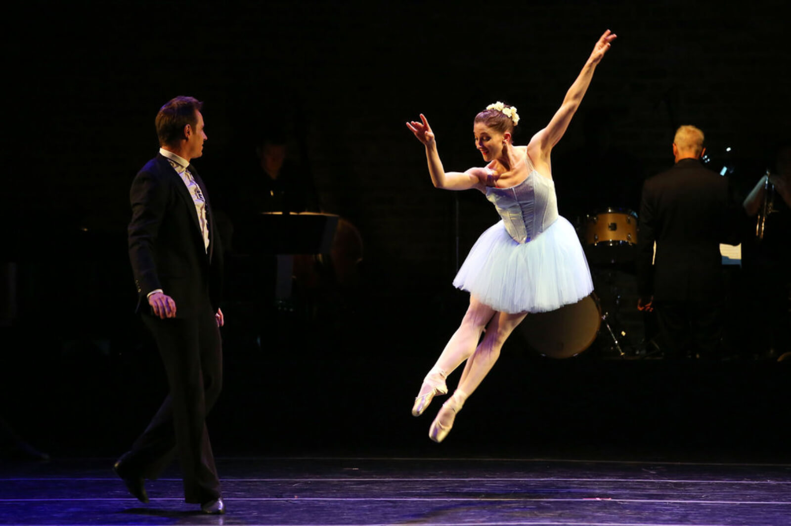 A female dancer in a blue tutu leaps toward her dance partner in a suit.
