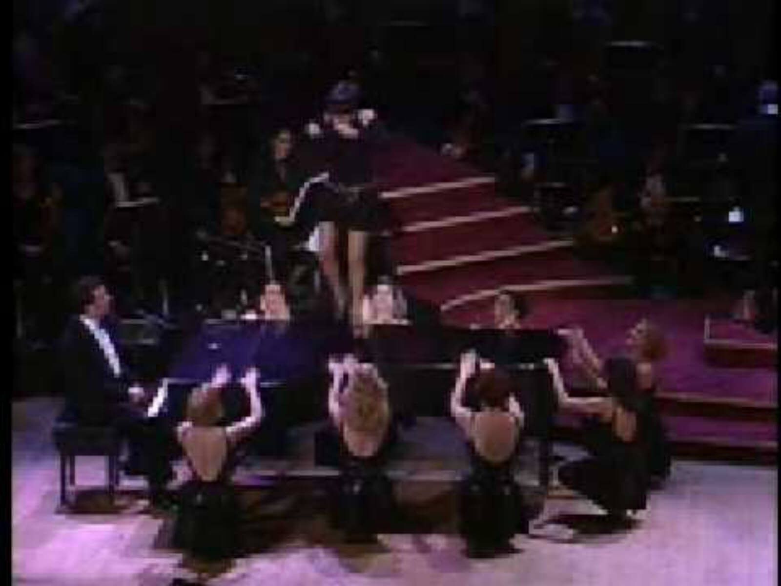 Liza Minnelli, in a beautiful black dress on top of a grand piano, sings and dances for the audience. She is with the female ensemble performing "Back in Business".